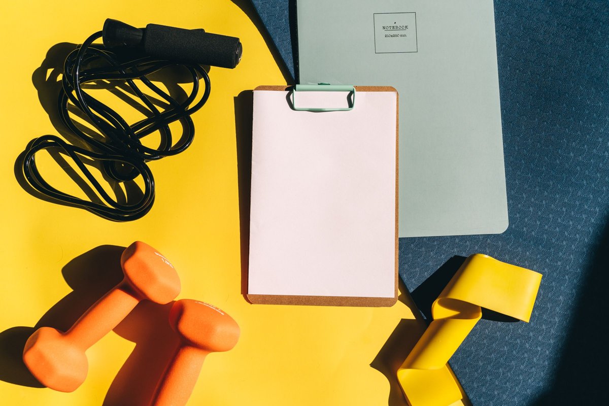 Dumbbells, jump rope, resistance band and a blank clipboard on a yellow and blue background