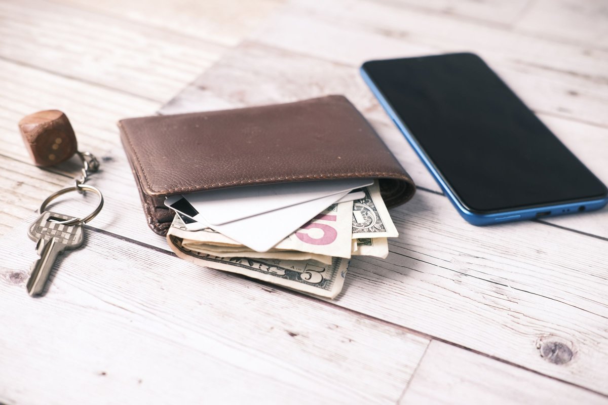 Brown wallet with cards and cash next to a smartphone and keys on a white wooden table